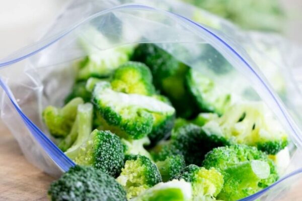 A person using the water displacement method to remove air from a hermetic freezer bag filled with bright green broccoli florets